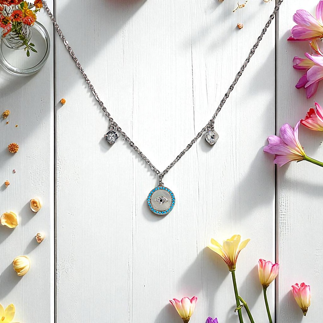 The Vellura Jewellery Evil Eye Pendant Necklace, crafted in sterling silver and featuring three pendants—one a turquoise circle—is displayed on a white wooden surface with colorful flowers and decorative glass vases.
