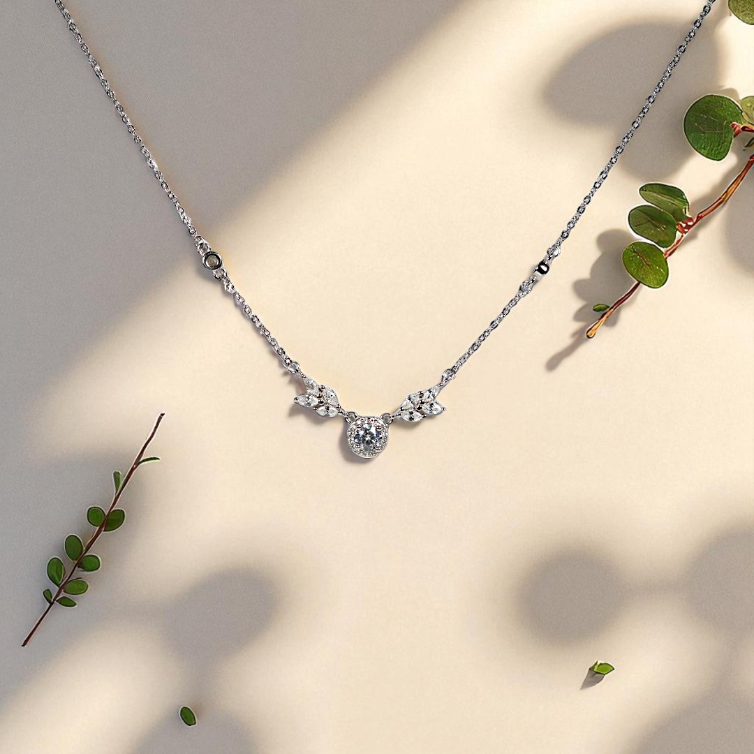 The Sterling Silver Pendent by Vellura Jewellery features delicate leaf and circle pendants, displayed on a beige surface with green sprigs and soft leaf shadows.